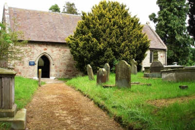 A view along the path to the south door of the church. The church is partially hidden by a large yew and there are gravestones and tombs in the churchyard in the foreground.
